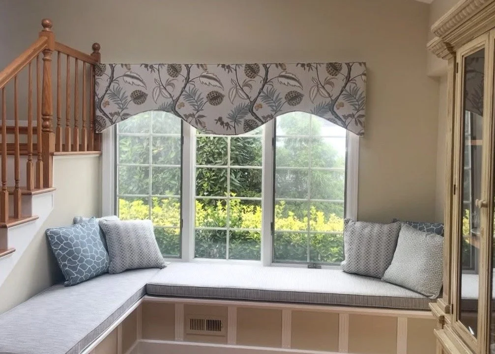 Built-in window seat beneath three large windows with floral valance, decorated with blue and white patterned pillows, next to a wooden staircase and a beige cabinet.
