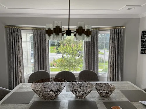 Dining room with a white table, three decorative wire baskets, gray placemats, and a modern chandelier. Large windows with patterned curtains and a view of greenery outside.