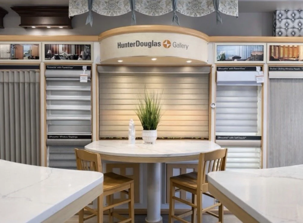 Display of Hunter Douglas window shades and blinds in a showroom with a round table, wooden chairs, and a potted plant.
