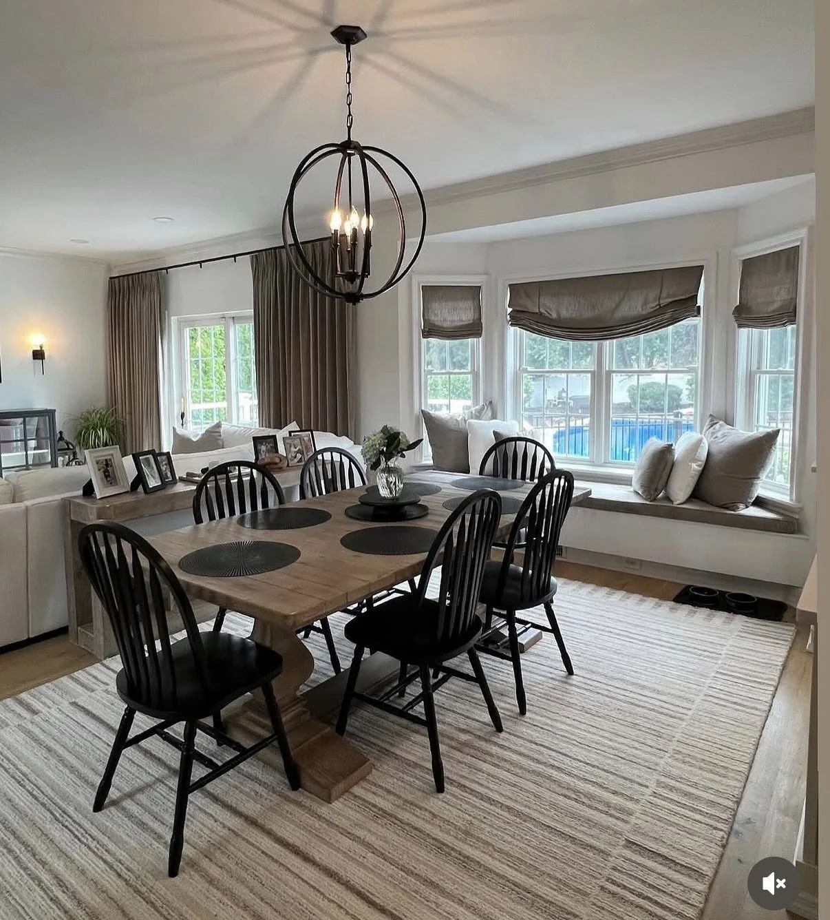 A dining room with a wooden table, black chairs, and a chandelier. There is a window seat with cushions and a large window with blinds. A rug is on the floor.