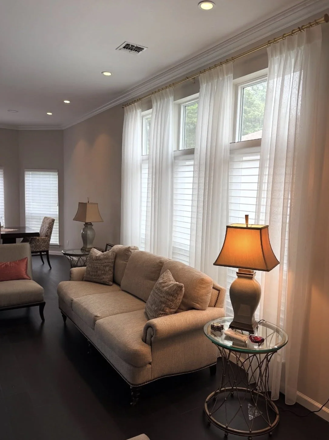 Living room with beige sofa, decorated pillows, side table with a lamp, and large windows with white curtains.