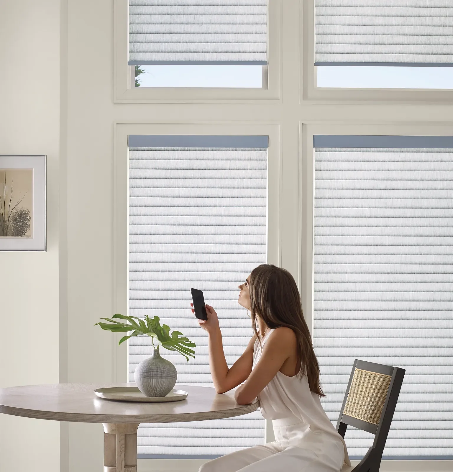 Woman sitting at a round dining table in front of large windows with white blinds, holding a smartphone with a plant in a gray vase on the table.