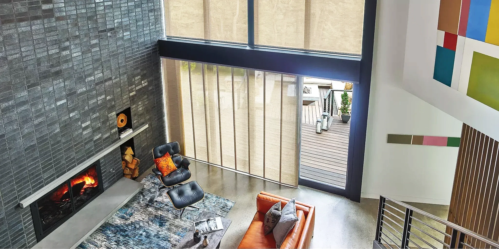 Modern living room with a fireplace, black leather chair, patterned rug, orange sofa, and large glass sliding door leading to an outdoor patio with potted plants.