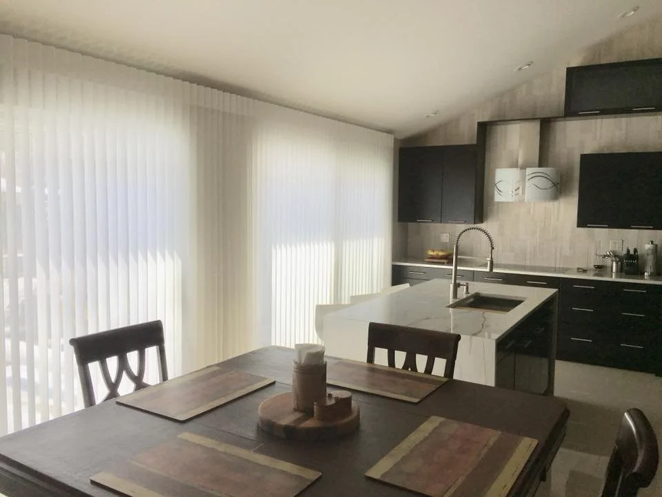 Modern kitchen with black cabinets, marble island, stainless steel sink, and floor-to-ceiling window with vertical blinds.