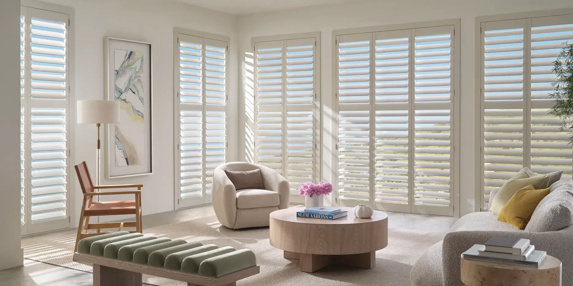 Bright living room with large windows covered in white plantation shutters, beige sofa with yellow and gray pillows, round wooden coffee table with pink flowers and books, a white armchair with a brown pillow, a wooden chair with pink cushion, a floor lamp, and abstract artwork on the wall.