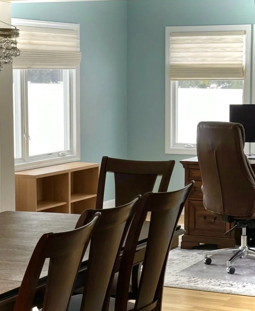 A bright dining room with two windows, light blue walls, a wooden dining table with chairs, a wooden cabinet, an office desk, and a brown office chair.