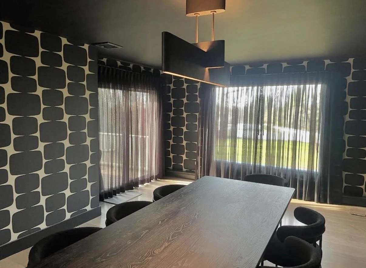 Interior of a modern conference room with a long wooden table and black chairs, patterned wallpaper on the walls, and large windows with sheer curtains allowing natural light.