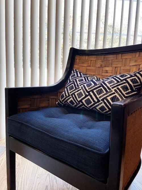 A wooden armchair with woven back and sides, dark frame, navy cushion, and a geometric patterned pillow, placed in front of vertical blinds.