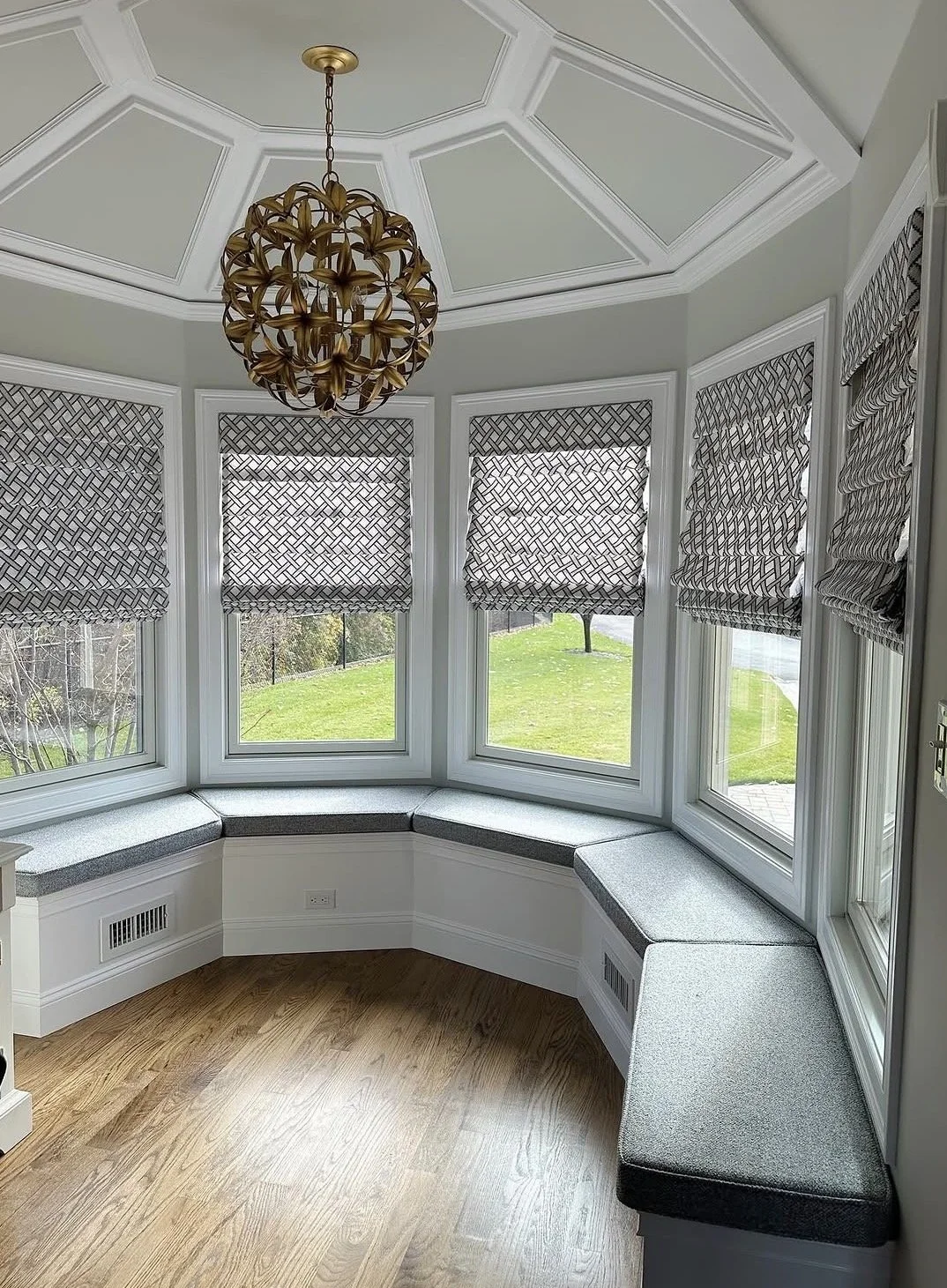 A cozy window nook with a built-in cushioned bench surrounding a bay of windows in a bright room with a decorative ceiling and a gold chandelier.