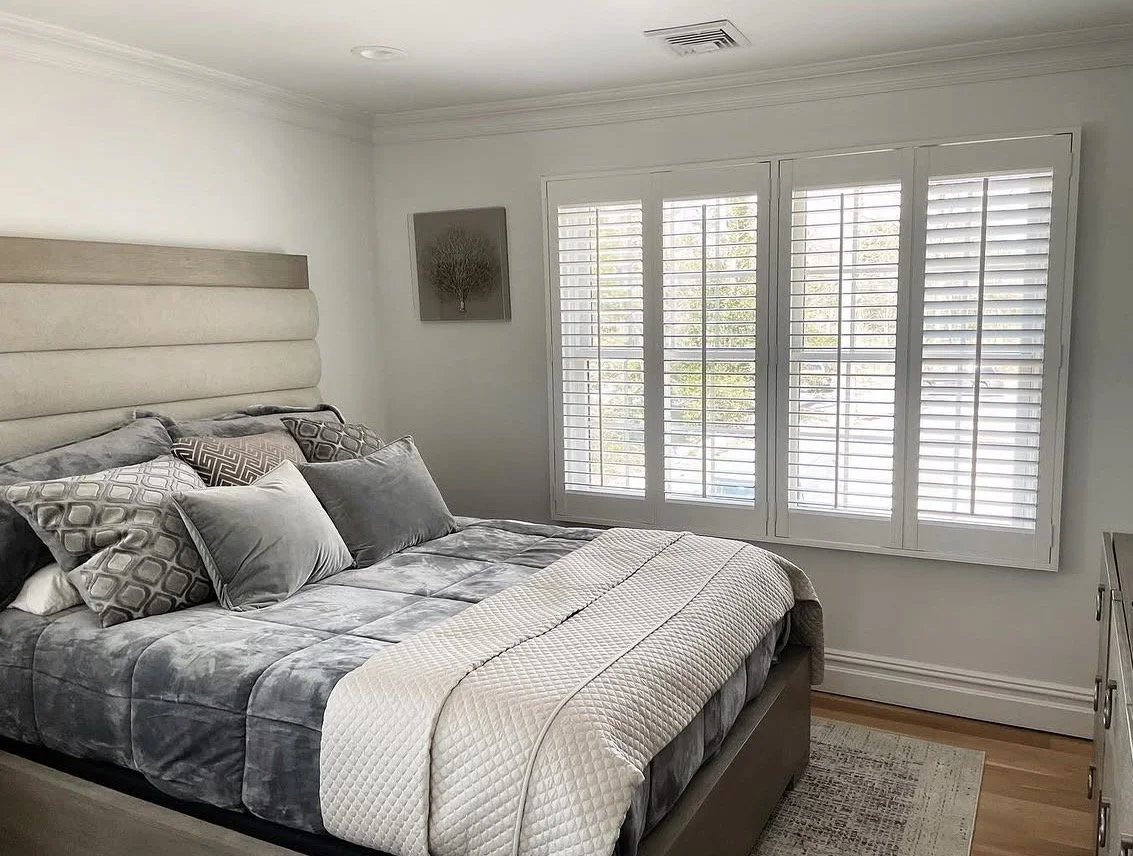 A neatly made bed with gray and white bedding and multiple pillows in a bedroom with large window shutters letting in natural light.