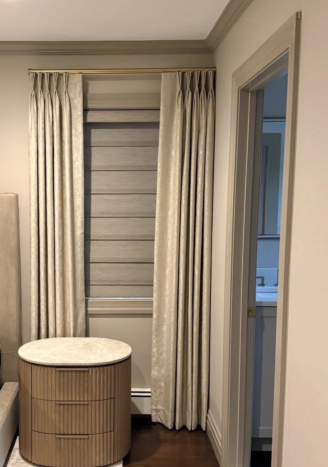 Interior corner with beige curtains, a small oval wooden side table with a marble top, and a partial view of a bathroom through a doorway.