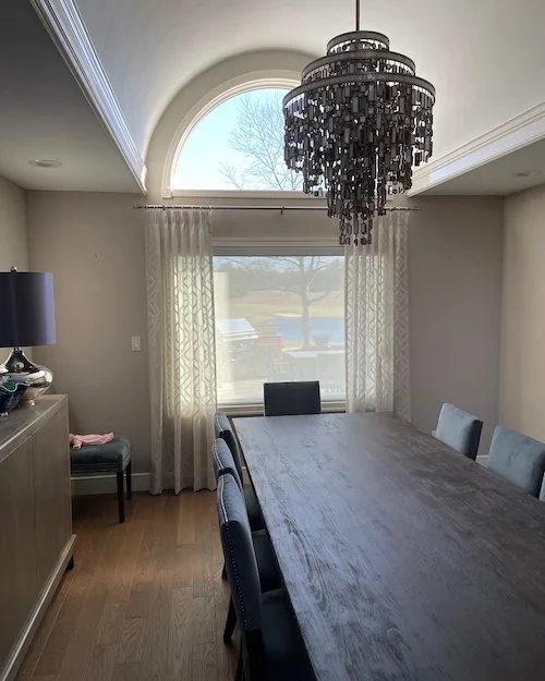 A dining room with a wooden table, six upholstered chairs, a large window with curtains, and a black chandelier hanging from the ceiling.