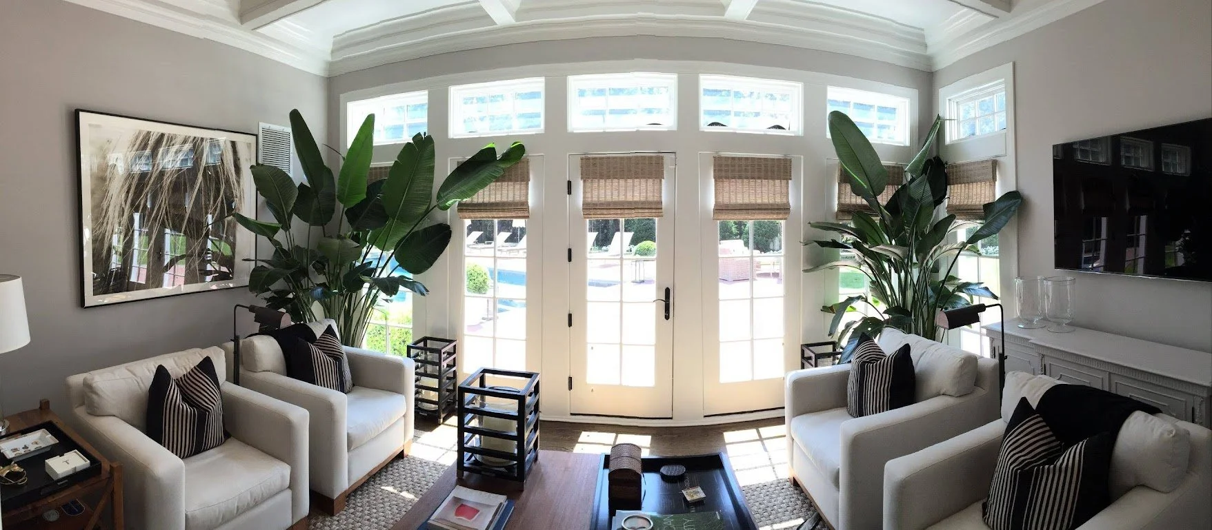 Bright living room with white sofas, black and white striped pillows, large green plants, a wall-mounted TV, and a front door with glass panes and woven blinds, allowing natural light to fill the space.