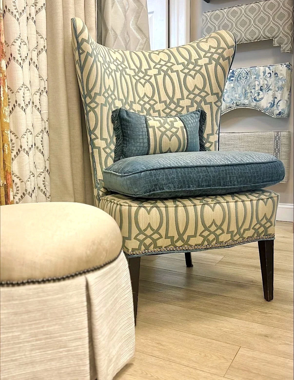 A patterned armchair with a high back, blue cushion, and dark wooden legs, surrounded by curtains, wall art, and a round beige ottoman.