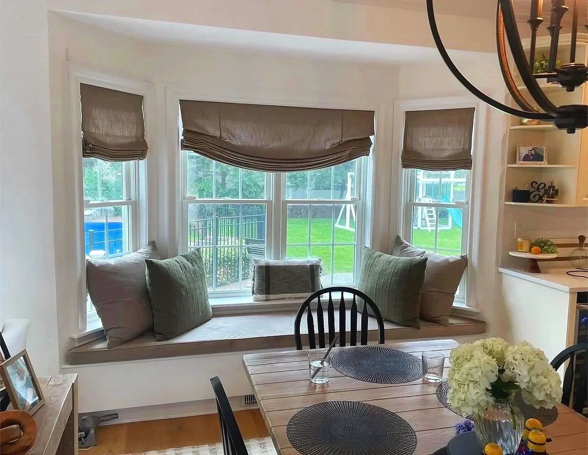 A breakfast nook with a large window seat, green and beige pillows, and three windows with beige Roman shades, overlooking a backyard with green grass and a blue slide.