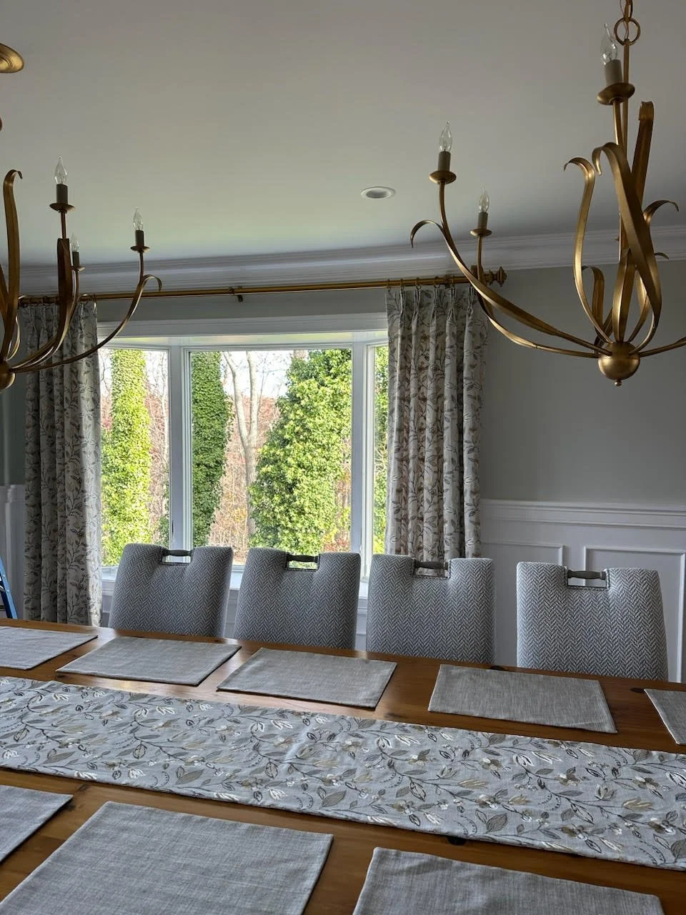 A dining room with a wooden table, six upholstered chairs, large window with floral curtains, and two gold chandeliers hanging from the ceiling.