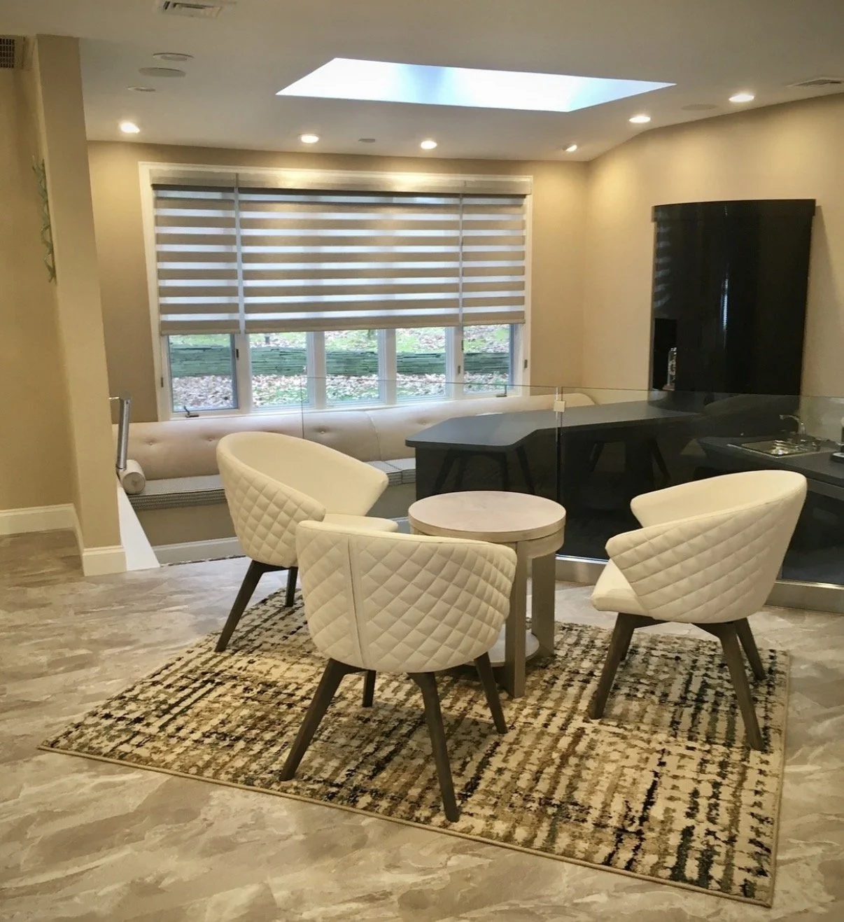 A cozy dining area with a round table, three quilted cream-colored chairs on a patterned rug, large window with horizontal blinds, and a skylight overhead in a modern room.