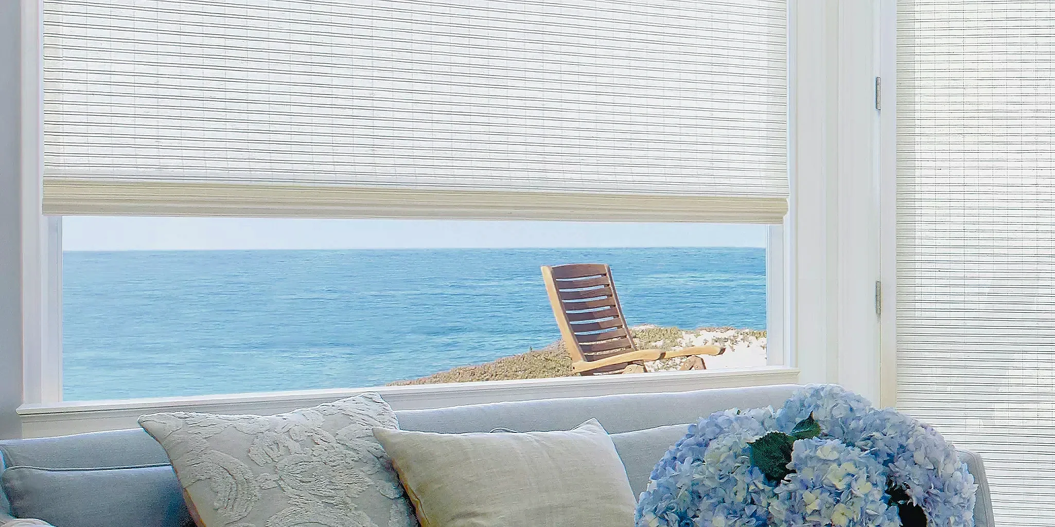 View of the beach and ocean through an open window with blinds. A wooden lounge chair on the sand outside.
