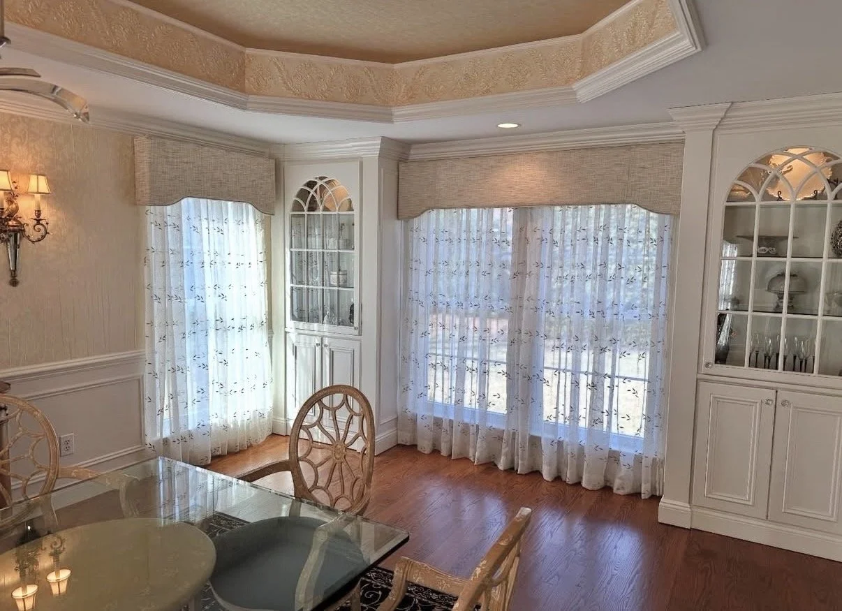 Dining room with large bay windows covered with sheer curtains and valances, wooden dining table with glass top, and matching wooden chairs with decorative backs. Built-in white cabinets display dishes and decorative items. Hardwood floor and wall sc