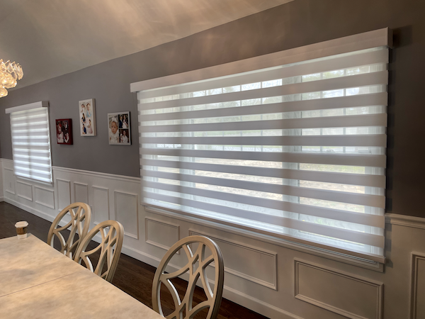 Modern dining room with large horizontal striped window blinds, gray wall with white wainscoting, framed photos, and beige chairs around a marble table.