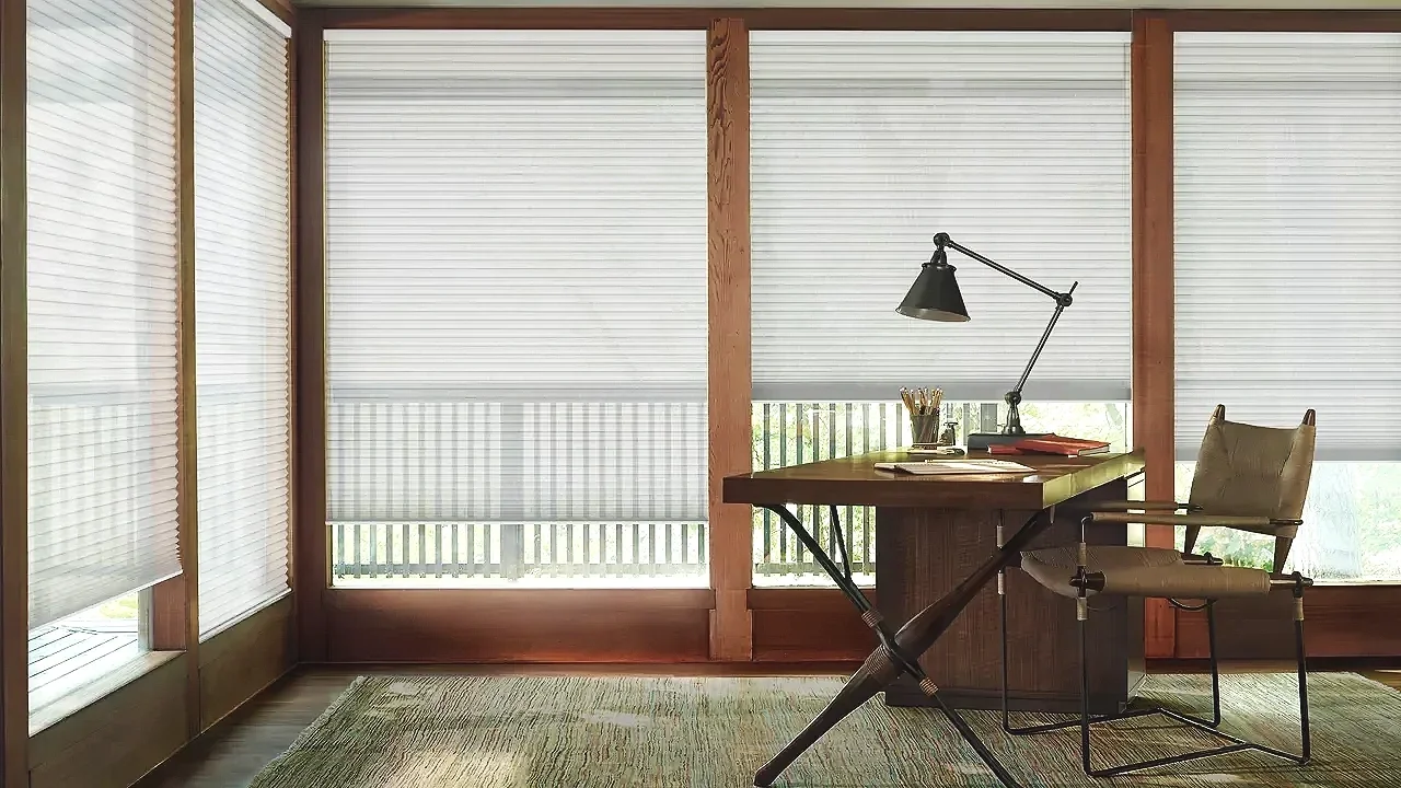 Home office with large windows covered by blinds, wooden desk with a black adjustable desk lamp, chair, and some office supplies