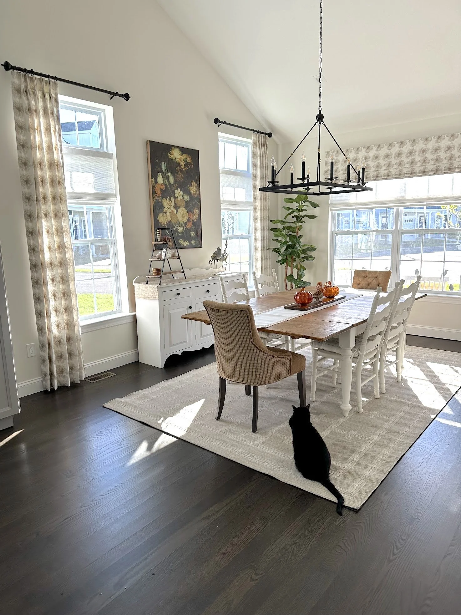 Bright dining room with a wooden table, white chairs, a beige armchair, and a black cat sitting on a cream area rug. Large windows with patterned curtains, a white sideboard, wall art, and a black chandelier light fixture hanging from the ceiling.