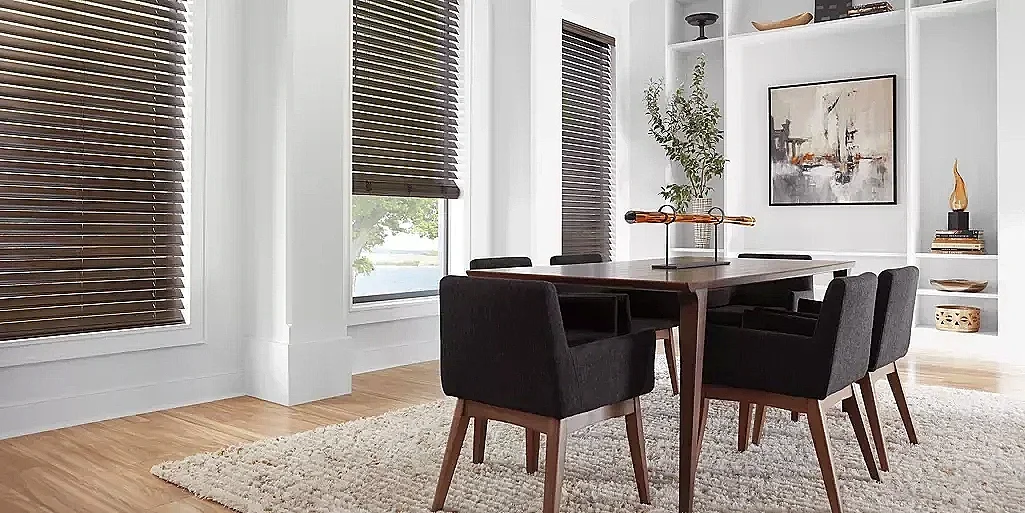 Dining room with a dark wood table, four black upholstered chairs, a beige rug, and large windows with brown blinds. Shelves and artwork are on the white walls.
