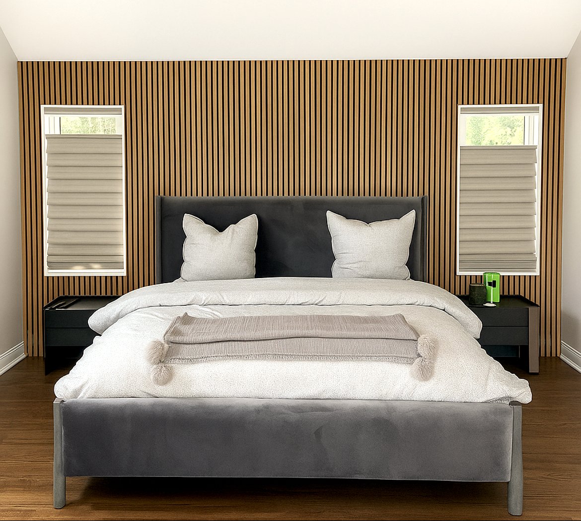 Modern bedroom with a grey upholstered bed, white bedding, two large pillows, and a beige throw blanket. There are black nightstands on each side with small lamps and a green device on the right nightstand. The wall behind the bed features vertical wooden slats flanked by two windows with beige blinds, allowing natural light into the room.