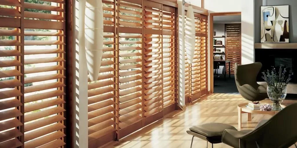 Sunlight streams through wooden slat window blinds in a bright living room, casting striped shadows on the wooden floor.