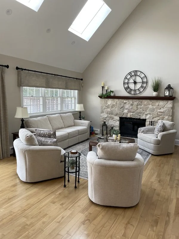 A cozy living room with high vaulted ceiling and skylights, featuring a stone fireplace with a wooden mantle, a large clock, and decorative items. The room has a large sofa, two armchairs, side tables, and lamps, with hardwood flooring and a window w
