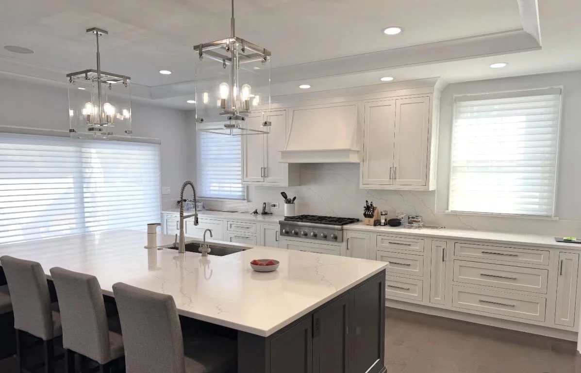 Modern white kitchen with island, pendant and ceiling lights, white cabinets, marble backsplash, and windows with blinds.