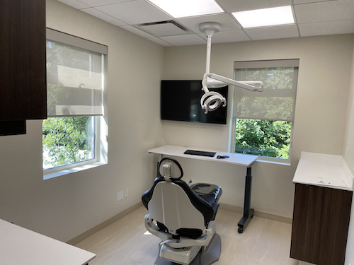A medical examination room with two windows, a mounted monitor, an adjustable chair, a ceiling-mounted medical light, and a white desk.