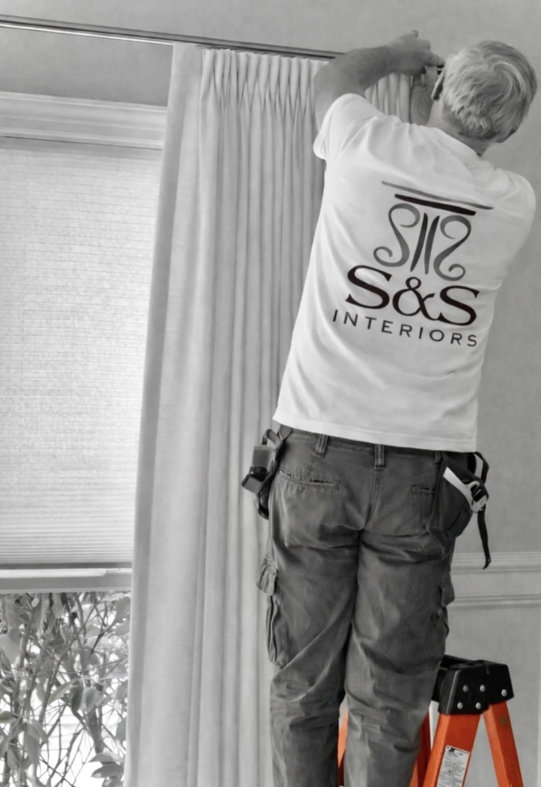 Worker adjusting curtain on window using a ladder, wearing a S&S Interiors t-shirt and tool belt, with a window and curtains in the background.