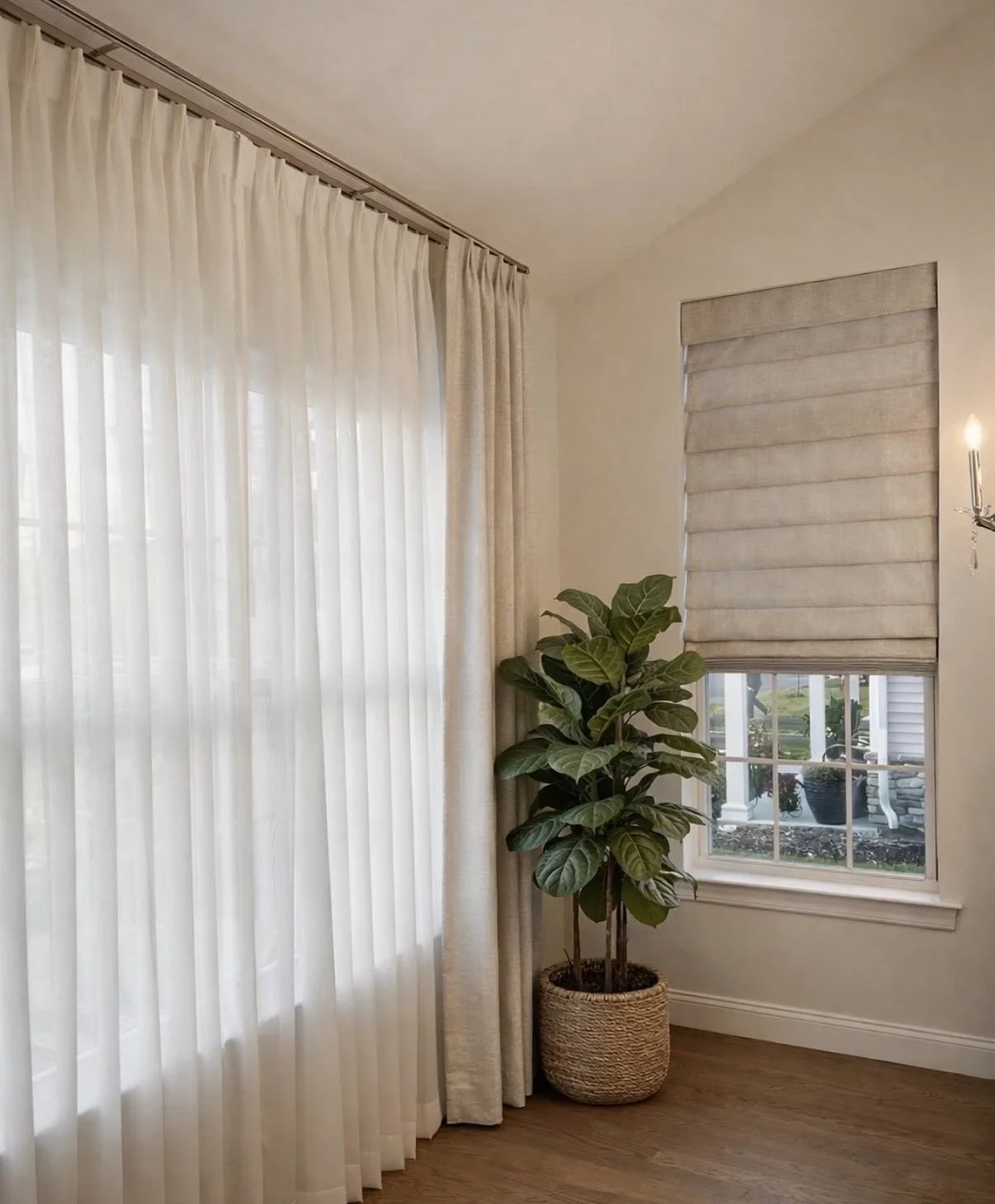 Interior of a living room corner with beige curtains, a fiddle leaf fig plant in a woven basket, and a window with a beige Roman shade.