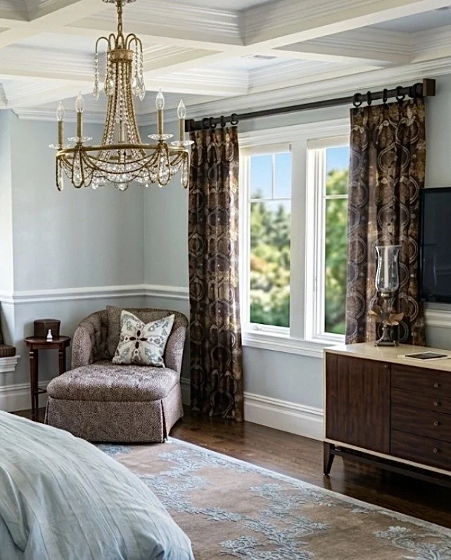A bedroom corner with a beige upholstered armchair, floral throw pillow, window with patterned curtains, a wooden side table, a crystal chandelier, a wooden dresser with a glass vase, a flat-screen TV, and a light-colored rug on dark hardwood floors.