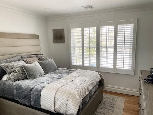 A neatly made bed with gray and white bedding in a bright bedroom with large window shutters, a painting on the wall, and a dresser.