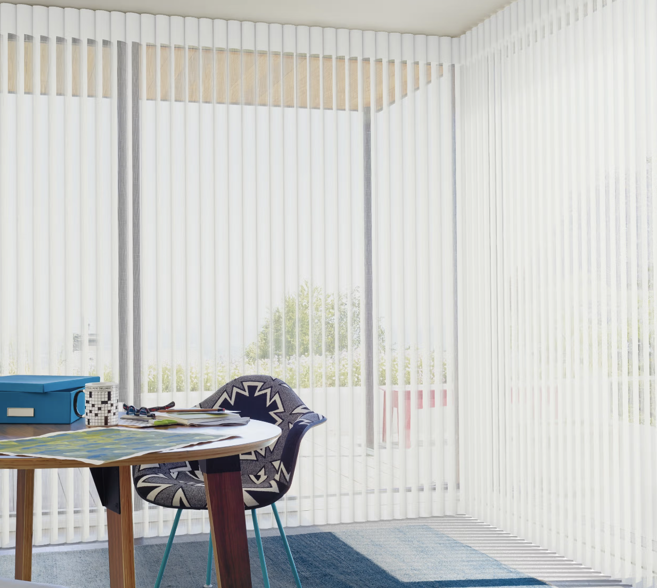 Bright office space with large windows covered by vertical blinds, a round wooden table with papers, books, and a blue storage box, and a modern black and white patterned chair.