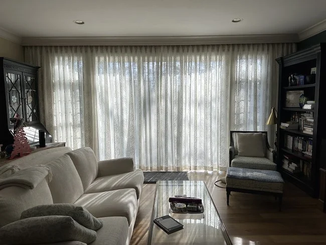 Living room with cream-colored sofa, armchair with cushion, glass coffee table, and large window with sheer curtains. There are bookshelves and a lamp in the background.