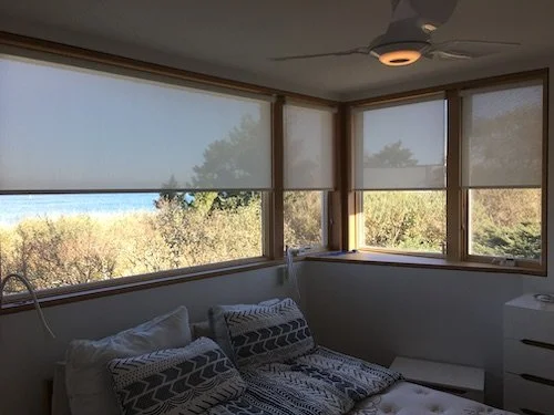 A bedroom with large windows showing trees and a view of the sky outside, a bed with patterned pillows, a ceiling fan, and a white dresser.
