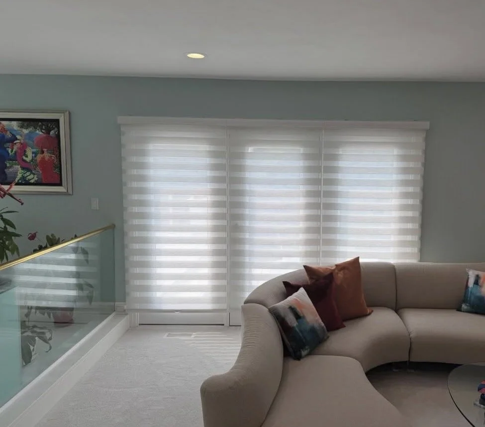 Living room with a beige sectional sofa, colorful throw pillows, white horizontal-striped window blinds, and a framed colorful picture on a light blue wall