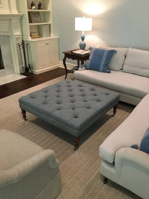 A cozy living room with a white sofa, a matching white armchair, and a large blue tufted ottoman in the center. There is a side table with a lamp and a bookshelf on the wall.