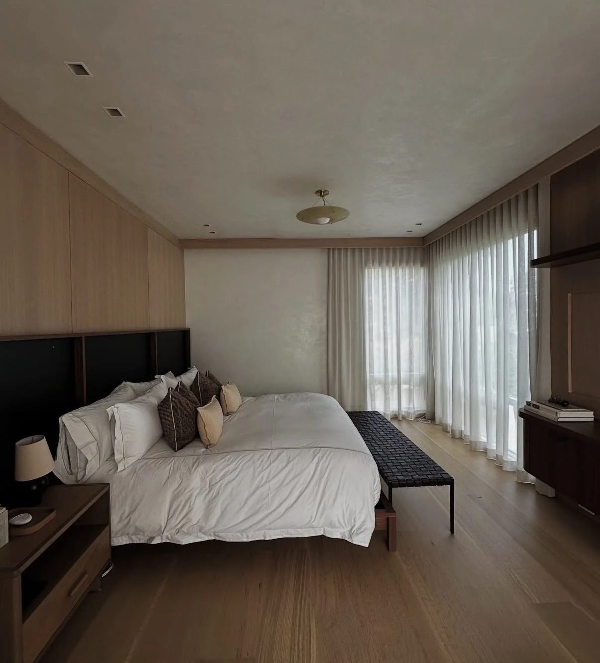 A bedroom with a large bed, dark headboard, white bed linens, and multiple pillows. There is a small side table with a lamp next to the bed, and a black bench at the foot of the bed. Floor-to-ceiling windows with sheer curtains let in natural light, 