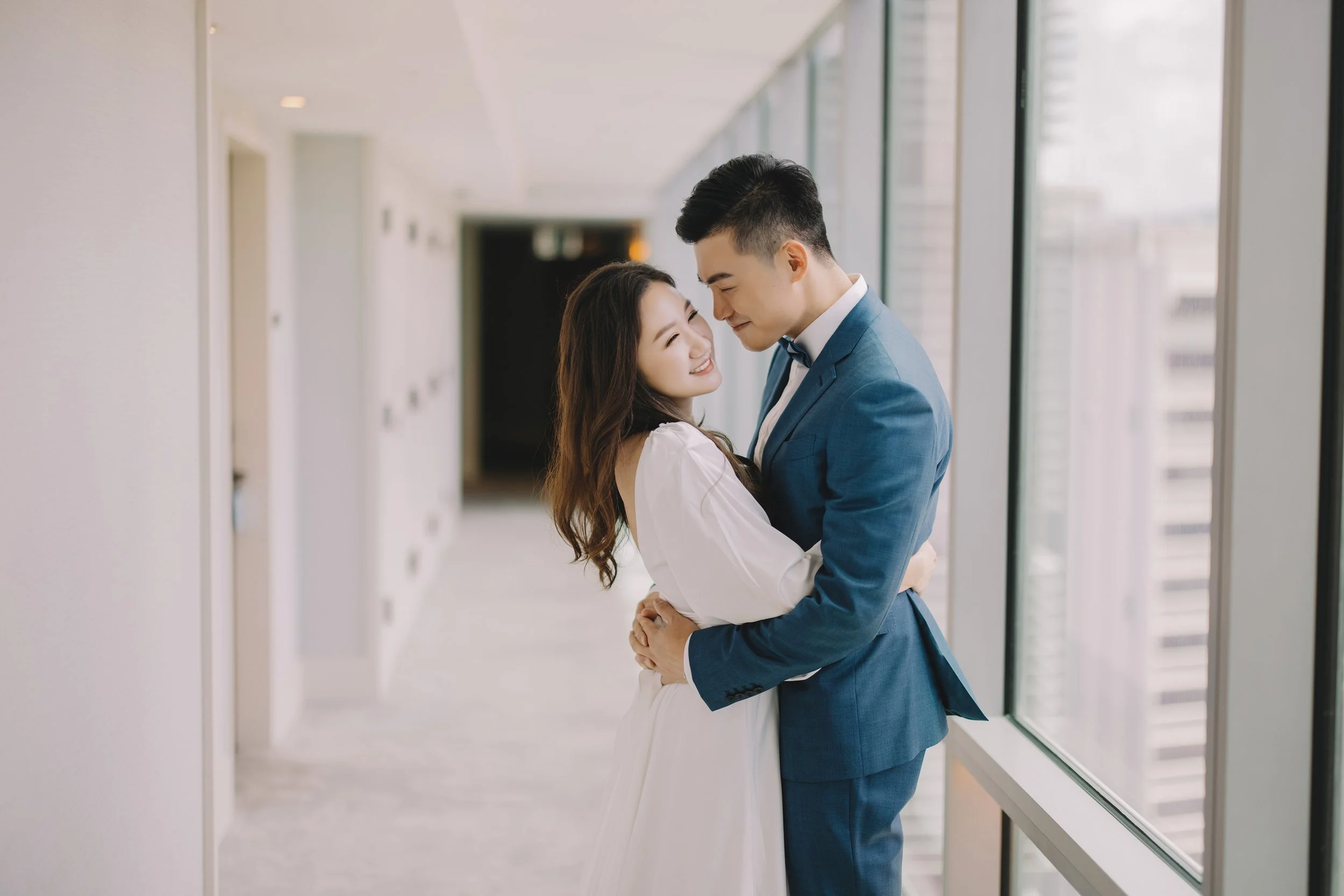 A couple dressed in formal attire sharing an affectionate moment by a large window in a bright, modern hallway.