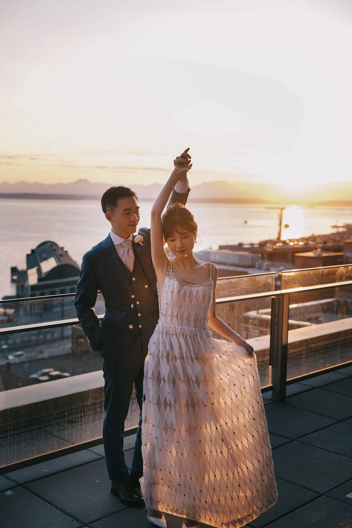 A couple dressed in formal attire enjoying a dance on a rooftop balcony overlooking a body of water during sunset.
