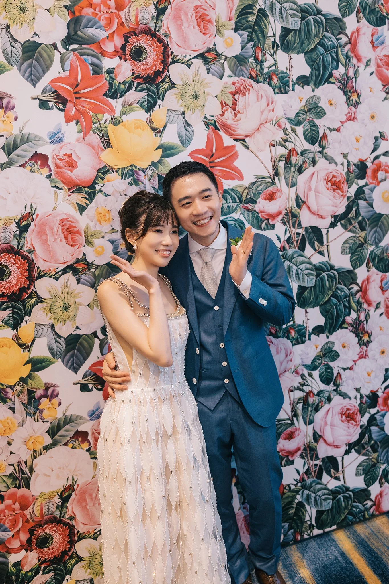 A smiling couple posing in front of a colorful floral wallpaper at a celebration, with the woman wearing a white dress and the man in a blue suit.