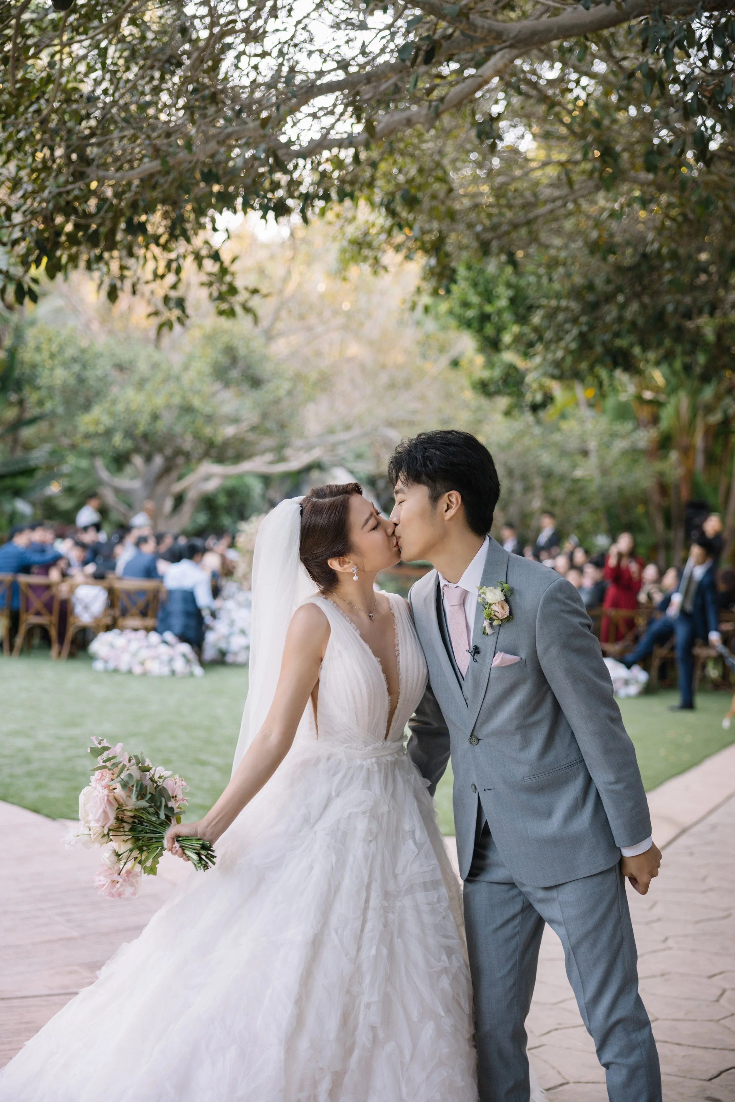 A bride and groom kiss during their wedding ceremony outdoors, with guests seated and trees in the background.