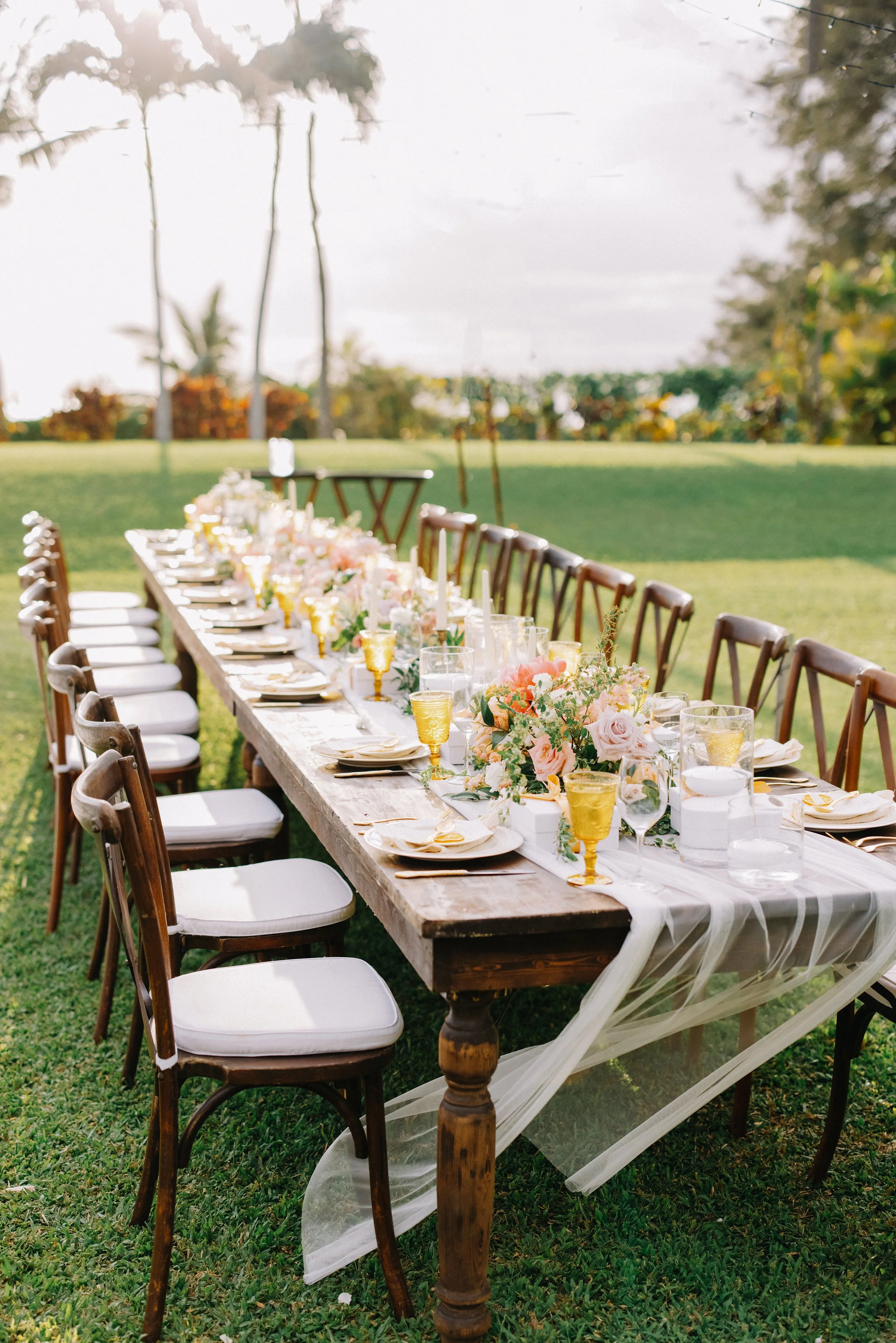 Long wooden outdoor dining table decorated with pink and white flowers, gold-rimmed glasses, white plates, and white napkins, set up on a grassy lawn with chairs around it, under a partly cloudy sky.