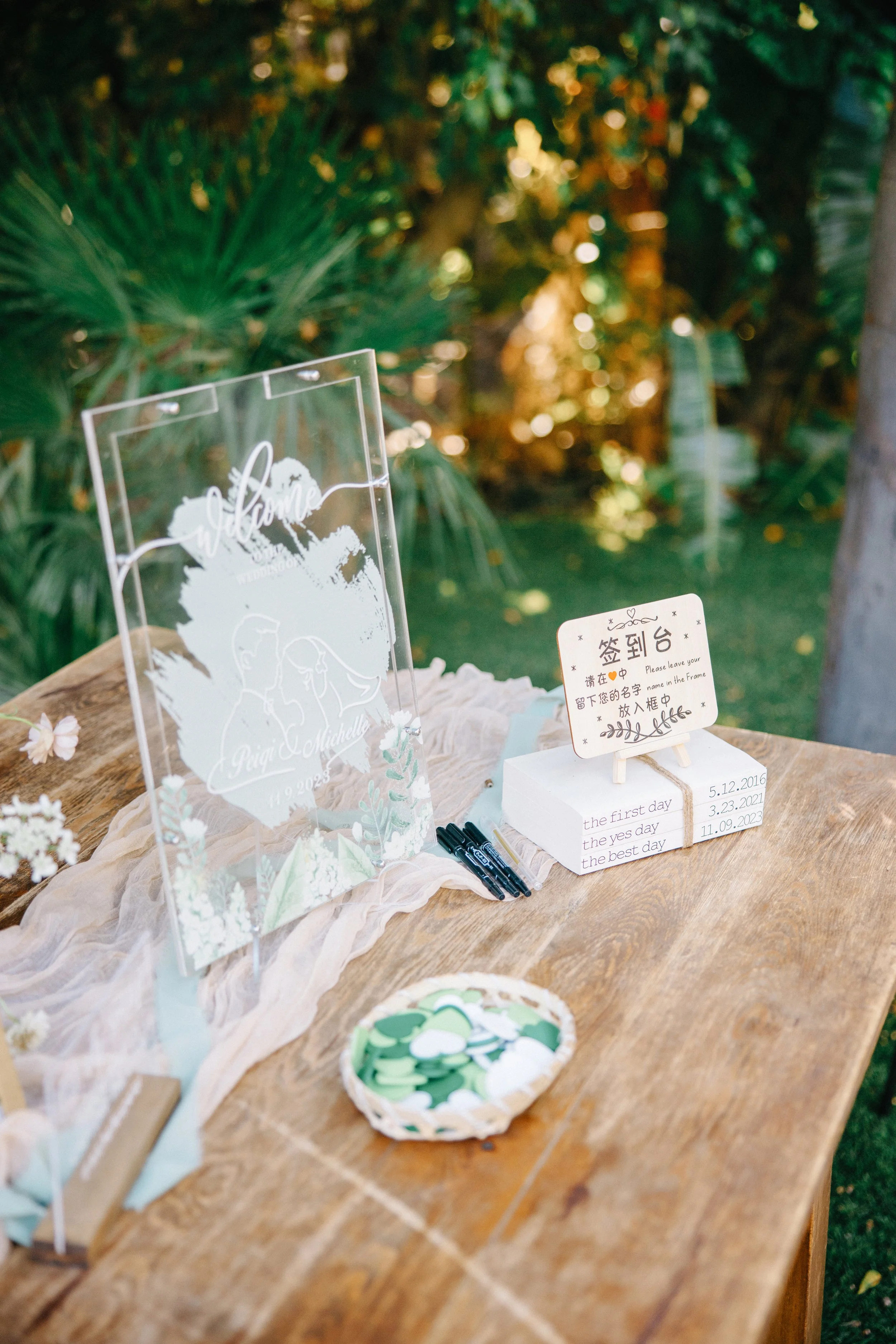 Wedding sign with illustration of a couple, a sign-in sheet with dates, and a basket of green and white paper hearts on a wooden table outdoors surrounded by greenery.