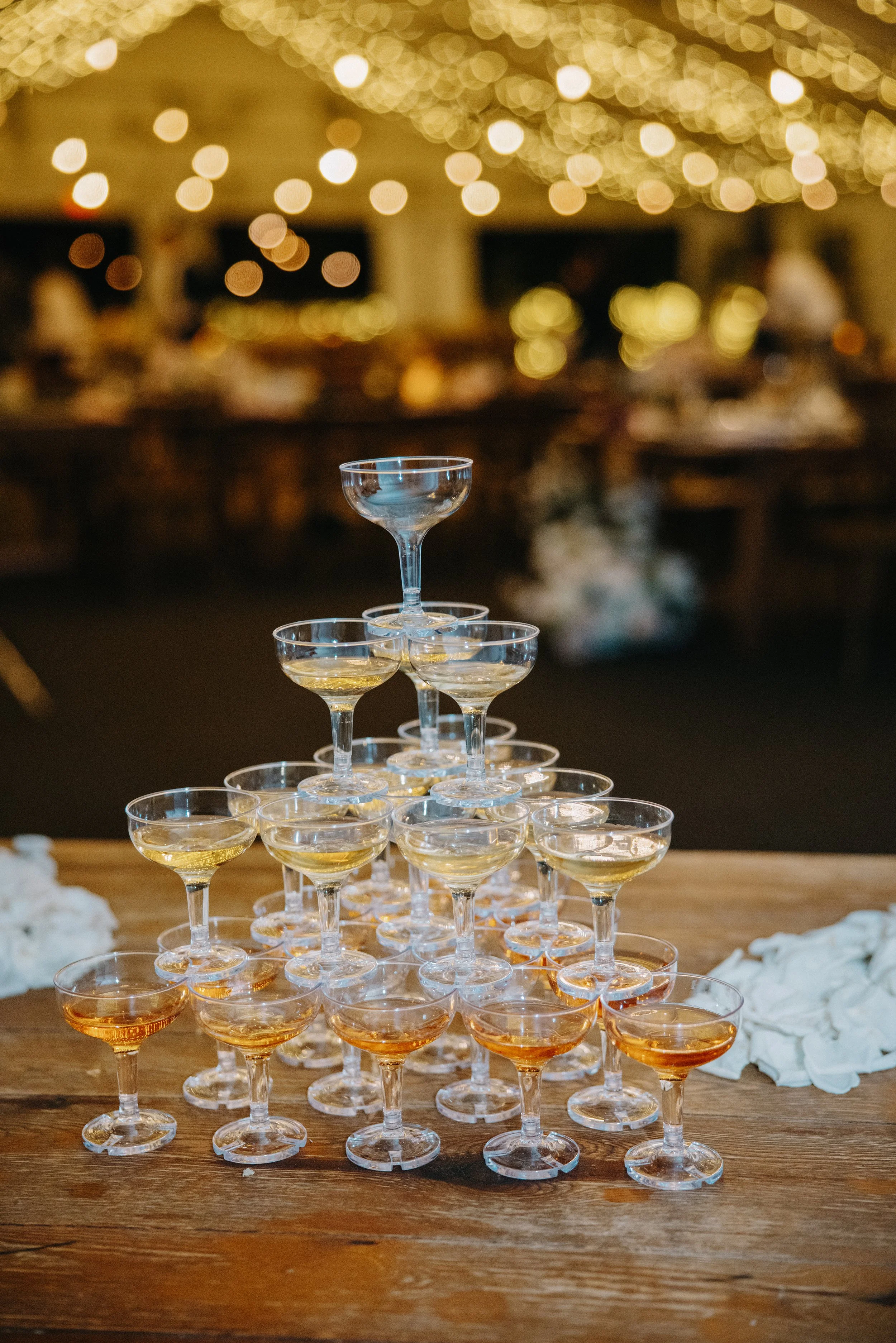 A pyramid of champagne glasses filled with a bubbly drink at a celebration event, with warm string lights and a blurred lively background.