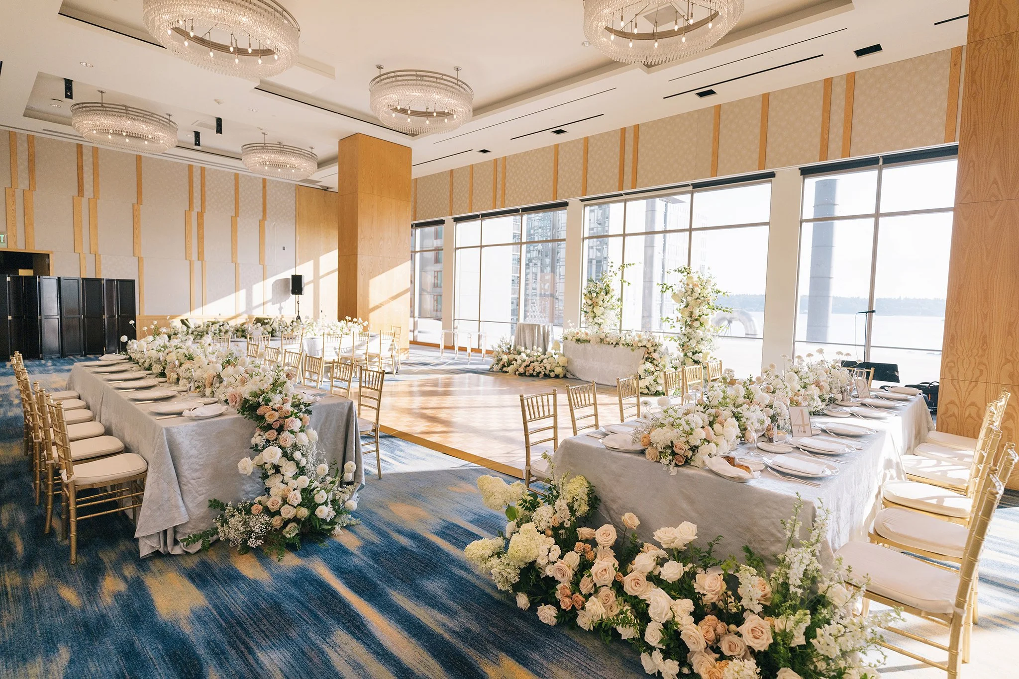 Elegant indoor wedding reception setup with long tables decorated with white and blush flower arrangements, gold chairs, and large windows providing natural light and a view outside.